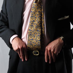 Pode incluir: Um conjunto formal com uma gravata estampada em azul marinho e dourado, uma camisa rosa claro e um blazer preto. O traje inclui um cinto preto com fivela dourada. O visual geral é clássico e profissional.