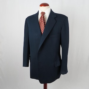 May include: A navy blue blazer on a mannequin. The jacket features a classic two-button design with notched lapels and two front pockets. A white dress shirt and a red patterned tie are visible. The blazer is a standard fit.