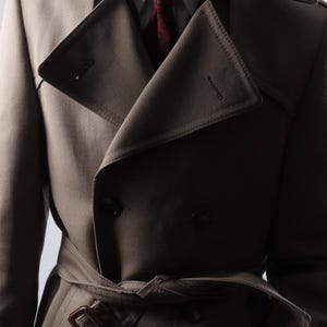 Puede incluir: Un abrigo tipo gabardina de color beige con diseño de doble botonadura, cinturón y cuello con solapas. El abrigo se lleva sobre una camisa blanca y una corbata roja estampada. El abrigo tiene un aspecto clásico y a medida.