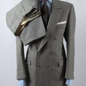 Puede incluir: Un traje cruzado de color gris con estampado de cuadros galeses y pantalones a juego colocados sobre el hombro. La chaqueta incluye un pañuelo de bolsillo blanco, con una camisa de rayas azul claro y blanca y una corbata oscura estampada.