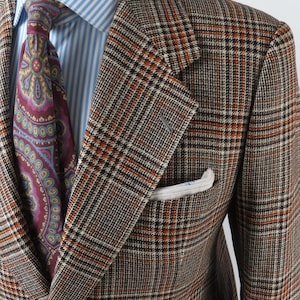 Puede incluir: Una americana de cuadros marrones, negros y naranjas sobre una camisa de vestir a rayas azul claro y blancas. Una corbata de color rojo oscuro con un estampado de cachemira multicolor está anudada al cuello y un pañuelo blanco en el bolsillo.