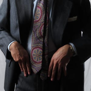 Pode incluir: Um fato cinzento-carvão composto por um casaco e calças a condizer, combinado com uma camisa de riscas finas azul-claras e brancas. Uma gravata larga apresenta um padrão paisley em vermelho escuro, azul e dourado. Um lenço branco no bolso.