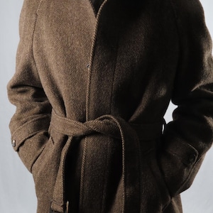 Puede incluir: Un abrigo marrón con estampado de espiga y cinturón a juego. El abrigo tiene un diseño clásico con cuello con solapas, puños abotonados y bolsillos. El cinturón está atado a la cintura. El abrigo es de tejido texturizado.