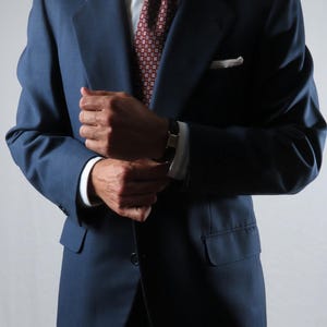 Puede incluir: Una americana azul marino sobre una camisa de vestir a rayas azul claro y blancas. Una corbata de color rojo oscuro con un pequeño patrón geométrico blanco está anudada al cuello. Un pañuelo blanco está metido en el bolsillo del pecho.