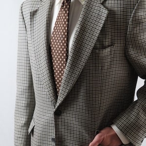 May include: A houndstooth patterned blazer in shades of gray and tan, paired with a white dress shirt and a brown tie with white polka dots. The blazer has a notched lapel and a single-button closure. The person is wearing the blazer with their hand in the pocket.