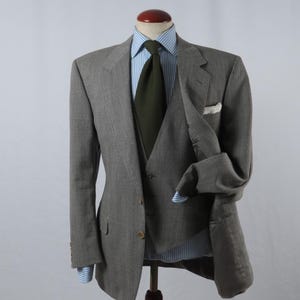 May include: A grey herringbone blazer worn over a matching vest and a light blue and white striped dress shirt. The ensemble includes a solid dark olive green necktie and a white pocket square tucked into the blazer's breast pocket.