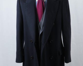 Op de afbeelding: Een marineblauwe dubbelbreasted overjas op een mannequin. De jas heeft een brede revers en een lange lengte. Een bordeauxrode stropdas en een grijs vest zijn eronder zichtbaar. Klassiek ontwerp.