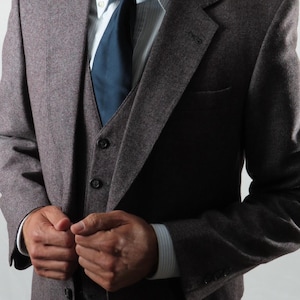 May include: A dark grey tweed three-piece suit, comprising a jacket, waistcoat, and trousers. Beneath the jacket, a light blue and white striped shirt is visible, paired with a dark blue tie.