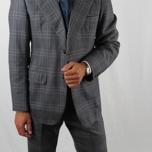 May include: A grey plaid suit, white shirt, and navy blue tie. The jacket has two buttons and a chest pocket. A brown wristwatch is visible. The suit is a classic formal wear ensemble.