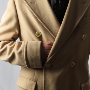 Peut inclure: Un pardessus beige à double boutonnage avec six boutons et un revers cranté. Le manteau a une coupe ajustée, une poche de chaque côté et un revers de manche. La personne porte une chemise blanche et une cravate à motifs.