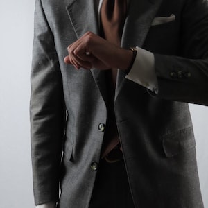 Könnte beinhalten: Ein anthrazitfarbener Blazer mit strukturierter Oberfläche, getragen über einem weißen Hemd mit Kragen und einer einfarbigen braunen Krawatte. Der Blazer hat ein steigendes Revers, zwei Knöpfe vorne und ein weißes Einstecktuch.