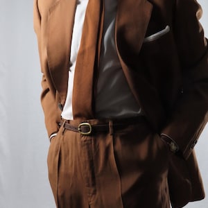 Puede incluir: Un traje marrón compuesto por chaqueta y pantalón a juego, combinado con una camisa blanca de cuello rígido y una corbata marrón texturizada. Se observa un cinturón de cuero marrón oscuro con hebilla de latón y un pañuelo blanco en el bolsillo.