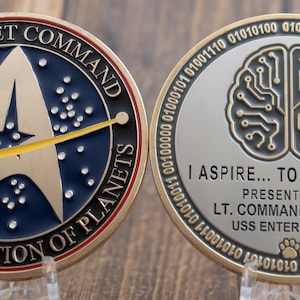 May include: A gold-coloured coin with two sides. The front side features the Starfleet Command logo with the text "Starfleet Command United Federation of Planets". The back side features a stylised brain with the text "I aspire... to be better. Presented by Lt. Commander Data USS Enterprise-D".