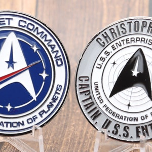 Peut inclure: Deux pièces de monnaie de défi en argent sur le thème de Star Trek. La première pièce présente le logo du Commandement de la Flotte stellaire avec un fond bleu et une étoile rouge et blanche. La deuxième pièce présente le texte "Christopher Pike" et le texte "U.S.S. Enterprise NCC-1701" avec une étoile noire et blanche.