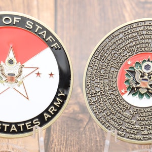 Puede incluir: Una moneda de color dorado con un diseño de estrella roja, blanca y azul en un lado y una lista de nombres de los Jefes de Estado Mayor del Ejército de los Estados Unidos en el otro lado. La moneda dice "Chief of Staff United States Army".
