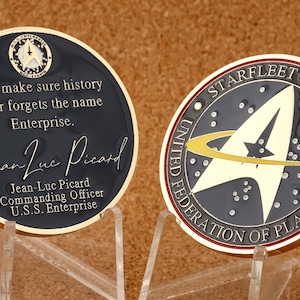 Op de afbeelding: Een gouden munt met een zwarte achtergrond en een gouden Starfleet-embleem. De tekst "STARFLEET COMMAND UNITED FEDERATION OF PLANETS" staat rond het embleem. De andere kant van de munt heeft een zwarte achtergrond met gouden tekst die "Let's make sure history never forgets the name Enterprise." en "Jean-Luc Picard" met een handtekening luidt. De tekst "Jean-Luc Picard Commanding Officer U.S.S. Enterprise" is ook inbegrepen.