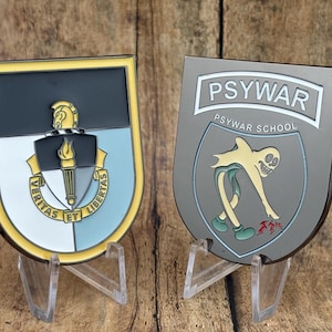 May include: Two metal military-style plaques on clear stands. One is shield-shaped with a black, gold, and light blue design, a torch, and the words "VERITAS ET LIBERTAS." The other is gray with "PSYWAR SCHOOL" and a cartoon figure.