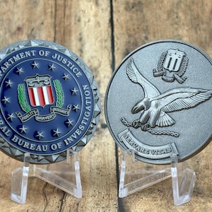 May include: Two metal coins displayed on clear stands. One coin features the text "FEDERAL BUREAU OF INVESTIGATION" and "DEPARTMENT OF JUSTICE" around a blue center with a shield and stars. The other coin shows an eagle and the text "SERVARE VITAS".