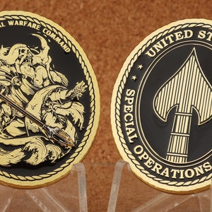 Puede incluir: Una moneda de color dorado con fondo negro. Un lado presenta una representación de una criatura marina mítica sosteniendo un tridente. El texto "NAVAL SPECIAL WARFARE COMMAND" está inscrito alrededor del borde. El otro lado presenta una punta de lanza dorada con el texto "UNITED STATES SPECIAL OPERATIONS COMMAND" inscrito alrededor del borde.