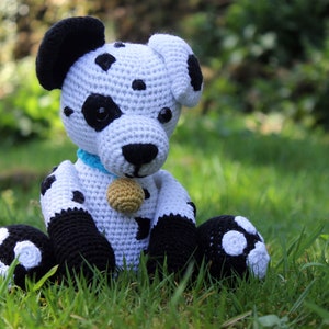 Puede incluir: Un juguete de peluche de dálmata tejido a crochet con manchas negras, sentado en la hierba verde. El juguete tiene un collar azul y una campanilla dorada.