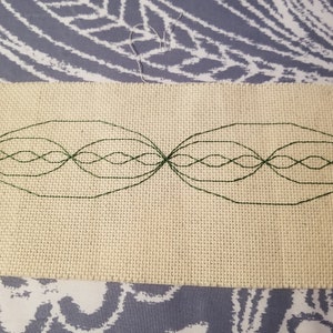 Puede incluir: Un diseño bordado verde sobre un fondo de tela beige. El diseño es una serie de óvalos superpuestos, creando un patrón ondulado.