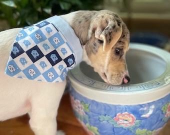 May include: A white and brown spotted puppy wearing a blue and white bandana with a repeated pattern of a blue and white logo. The puppy is looking at a blue and white ceramic pot with floral designs.