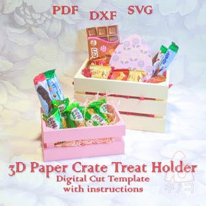 May include: A digital cut template for a 3D paper crate treat holder. The template includes instructions and is available in PDF, DXF, and SVG formats. The image shows two paper crates, one pink and one white, filled with various chocolates and candies.