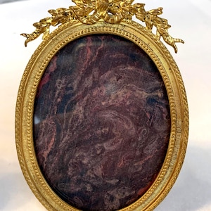 Può includere: Una cornice ovale con finitura in oro metallizzato e bordo testurizzato. La parte superiore presenta un motivo decorativo a fiocco e ghirlanda floreale. Il centro contiene un inserto scuro con venature nei toni del rosso, viola e nero.