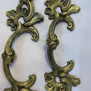May include: Two ornate gold-toned metal drawer pulls with a scrolled design. The pulls have a decorative floral pattern and a C-shaped handle.