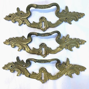 May include: Set of three antique brass drawer pulls with ornate, scroll-like designs. Each pull has a keyhole cutout and is in excellent condition.