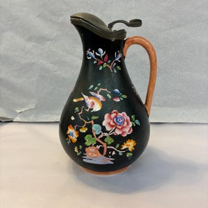 May include: A black ceramic pitcher with a metal lid and a light orange handle. The body features a hand-painted design of a colorful bird perched on a branch, surrounded by pink, orange, and blue flowers and green leaves against the dark surface.