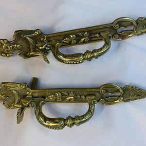 May include: Two ornate brass door handles with intricate floral designs and keyholes. The handles have a decorative, antique style.