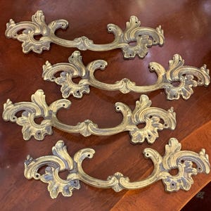 Puede incluir: Cuatro tiradores de cajón de latón adornados con un diseño de desplazamiento. Los tiradores tienen un acabado desgastado y antiguo.