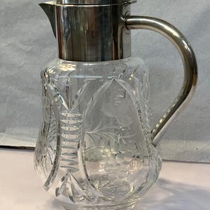 May include: A clear glass jug featuring intricate cut glass patterns and etched grape clusters. The vessel includes a polished silver-tone metal lid with a hinged spout cover and a matching curved metal handle attached to the neck.