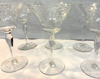 Op de afbeelding: Set heldere glazen martiniglazen. Elk glas heeft een conische kom, een slanke steel met een decoratief element en een ronde voet met geëtste details. De glazen zijn gerangschikt op een lichtgekleurd oppervlak.