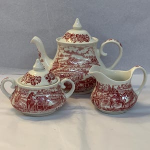 Könnte beinhalten: Ein weißes Keramik-Teeservice mit rotem Transferdruck-Design. Das Set beinhaltet eine Teekanne, eine Zuckerdose mit Deckel und einen Sahnekännchen. Das Design zeigt eine ländliche Szene mit Figuren, Pferden und Kutschen.