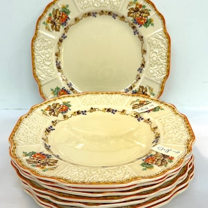 May include: A stack of six cream-colored ceramic plates with a raised floral design and a scalloped edge. The plates have a brown and orange floral pattern with a gold trim. The top plate has a handwritten note that says "Chip 7".