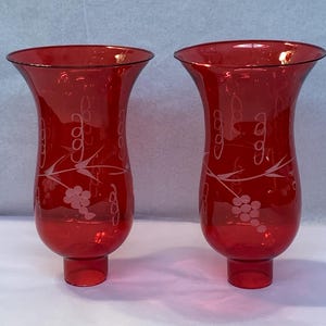 May include: A pair of ruby red glass candle holders. They have a flared top and a pedestal base. Each holder has an etched design of grapes and vines. The glass has a glossy finish. The design is elegant and the colour is striking.