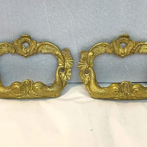 Puede incluir: Dos tiradores de cajones de color dorado con un diseño de sirena. Cada tirador tiene un marco decorativo enrollado y un diseño de cola de sirena en el centro.