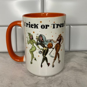 May include: White ceramic coffee mug with an orange rim and handle. The mug features a Halloween-themed design with three women dressed in costumes and the text "Trick or Treat".