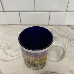 May include: White ceramic coffee mug with a blue rim and a colorful image of a building with the word "WENDEL" printed on the bottom.