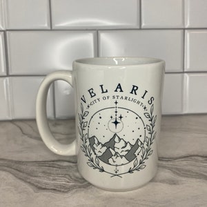 Puede incluir: Taza de cer&aacute;mica blanca con un dise&ntilde;o en negro y gris. El dise&ntilde;o presenta una cordillera, estrellas y el texto "Velaris, Ciudad de la Luz de las Estrellas".