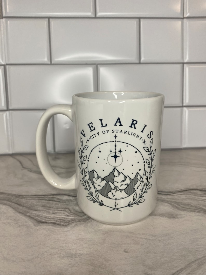 Puede incluir: Taza de cer&aacute;mica blanca con un dise&ntilde;o en negro y gris. El dise&ntilde;o presenta una cordillera, estrellas y el texto "Velaris, Ciudad de la Luz de las Estrellas".