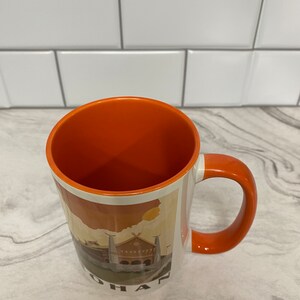 May include: Orange ceramic coffee mug with a white interior and a graphic of a building with the word "OHAN" at the bottom.