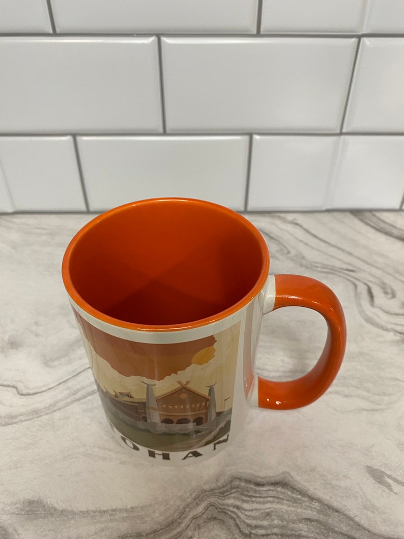 May include: Orange ceramic coffee mug with a white interior and a graphic of a building with the word "OHAN" at the bottom.
