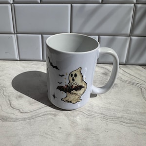 Peut inclure: Un mug en céramique blanche orné d'une illustration imprimée représentant un fantôme beige à l'expression surprise tenant une petite chauve-souris brune. Le motif comprend plusieurs petites chauves-souris noires et des étoiles à quatre branches.