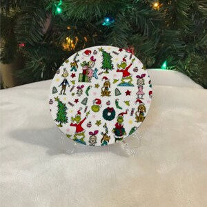 May include: A round white coaster with a Christmas-themed design featuring the Grinch and other characters from the book "How the Grinch Stole Christmas!" The coaster is decorated with green, red, and white colours and features a variety of festive elements, including Christmas trees, presents, and candy canes.