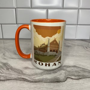 May include: A white ceramic mug with an orange rim and handle. The mug features a graphic of a building with a brown roof and a sunset sky. The text "ROHAN" is printed on the bottom of the mug.