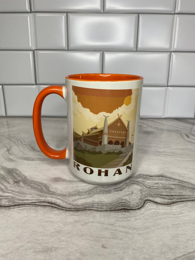 May include: A white ceramic mug with an orange rim and handle. The mug features a graphic of a building with a brown roof and a sunset sky. The text "ROHAN" is printed on the bottom of the mug.