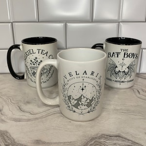 Puede incluir: Tres tazas de cer&aacute;mica blanca con asas negras y borde negro. Las tazas tienen dise&ntilde;os diferentes. La primera taza tiene una ilustraci&oacute;n en blanco y negro de una taza de t&eacute; y flores con el texto "RIEL TEA C" y "Tea In Prythian". La segunda taza tiene una ilustraci&oacute;n en blanco y negro de una cordillera con estrellas y el texto "VELARIS" y "CITY OF STARLIGHT". La tercera taza tiene una ilustraci&oacute;n en blanco y negro de un murci&eacute;lago con el texto "THE BAT BOYS" y "AIRIEL + RHYSAND + CASCAR".